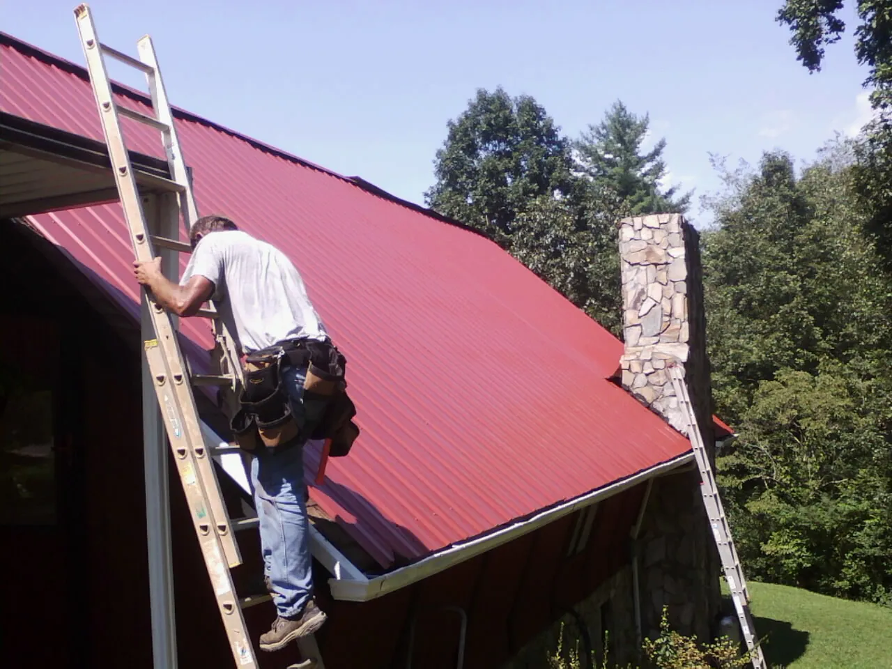 Roofer on a ladder beside a red metal roof and stone chimney — Soffit & Fascia Repair by Roofing Brothers in Eads